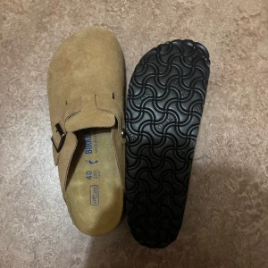 Birkenstock Boston - Birkenstock Boston sandaler i beige mocka med justerbart spänne och klassisk korksula. Skorna har en täckt tå och svart räfflad yttersula för extra grepp. Perfekta för chill vardagsstil och riktigt sköna att gå i.