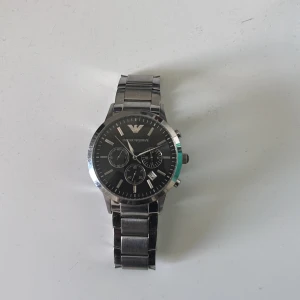 Emporio Armani silverfärgad klocka - Stilren klocka från Emporio Armani med svart urtavla och silverfärgat armband i metall. Klockan har tre mindre urtavlor, datumvisning och tydliga indexmarkeringar. Klassisk design med rund boett och länkarmband som ger en snygg och modern look. Klockan behöver nytt batteri då den stått länge utan användning. Ingår ej Box. 