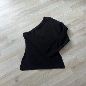 Svart oneshoulder topp från Zara - Stilren svart topp från Zara med oneshoulder-design och lång ärm på ena sidan. Toppens material är slätt och något stretchigt, vilket ger en snygg passform. Har knappast kommit till användning och det är varför jag säljer. Den är som ny och i jättebra skick. 