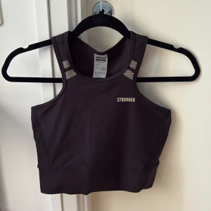Svart sporttopp från Stronger - Snygg svart sporttopp från Stronger med brottarrygg och diskreta meshdetaljer vid axlarna. Toppen är croppad och tillverkad i återvunnen polyester och elastan, vilket gör den stretchig och perfekt för träning. Logga framtill och baktill i guld. Aldrig använd, väldigt fina skick! 
