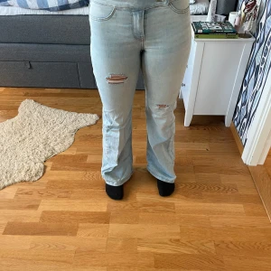 Ljusblå bootcut jeans med slitningar - Säljer ett par ljusblå jeans med bootcut-modell och snygga slitningar på båda benen. Navelhöjd på midjan och klassiska femficksdetaljer. Jeansen har en avslappnad vibe och är tillverkade i mjukt denimtyg. Perfekta för dig som gillar en trendig och bekväm look. Finns en lite prick av grön färg på vänster framsida men på pass liten att man inte märker den💕