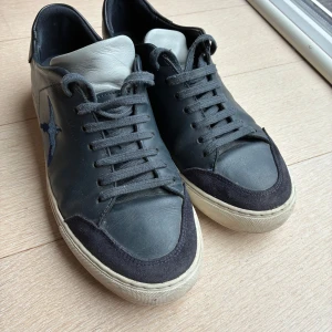 Blåa sneakers från Axel Arigato - Snygga blåa sneakers från Axel Arigato med vit sula och grå plös. Skorna har broderad blå fågel på sidan och detaljer i mocka vid tån. Klassisk rund tå och snörning framtill. Köpt på plock i slutet av 2024, använda ett fåtal gånger av mig. Säljer dem för de är för små för mig.