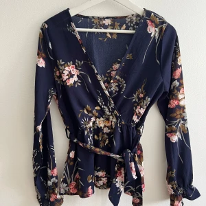 Blommig omlottblus med knytband - Mörkblå omlottblus med blommigt mönster i rosa, vitt och grönt. Blusen har långa ärmar med resår vid ärmslut och ett knytband i midjan som ger en snygg siluett. Passar bra för storlek XS och S.