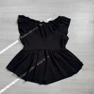 Svart volangblus med v-ringning - Supersöt svart blus med v-ringning och volangärmar. Modellen har en markerad midja och ett utställt, flowy avslut som ger en feminin siluett. Perfekt att styla med jeans eller kjol för en trendig look i storlek m men passar även S