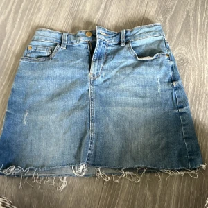 Blå jeanskjol från Lindex - Snygg ljusblå jeanskjol från Lindex med klassisk femficksdesign och rå, fransig fåll. Kjolen har bälteshällor, knapp och dragkedja framtill samt en bakficka. Perfekt för en avslappnad och trendig look.