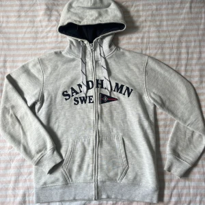 Ljusgrå Sandhamn zip hoodie - Ljusgrå SANDHAMN hoodie med dragkedja från SEA LIFE. Tröjan har broderad text, fickor och snören i huvan. Insidan av huvan är mörkblå och materialet är mjukt och skönt, perfekt för är kylig sommardag. Köpt för 799kr på Sandhamn. 