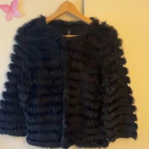 Svart pälsjacka med fluffig look - svart pälsjacka. Jackan har horisontella ränder i pälsen. Långärmad och utan synliga knappar. En knapp högst upp. Osäker på vart den kommer ifrån.