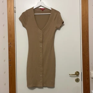 Beige ribbad kortklänning från Guess - Supersnygg beige ribbad kortklänning från Guess med v-ringning och korta ärmar. Klänningen har knappar hela vägen framtill med Guess-logga och en broderad logotyp nertill. Perfekt för en trendig och enkel look. Använd endast 1 gång!
