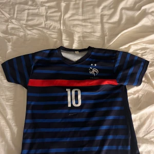 Frankrike Mbappé #10 fotbollströja - Snygg blå och svart randig Frankrike fotbollströja med röd detalj, nummer 10 och Mbappé på ryggen. Tröjan har korta ärmar, V-ringning och är gjord i ett lätt och ventilerande material. Perfekt för dig som vill bära landslagets färger och hylla Mbappé.