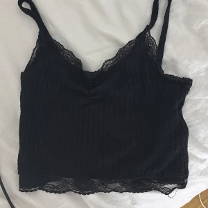 Svart ribbad linnetopp med spets - Svart croppad linnetopp från new yorker i storlek XXS. Toppen har tunna axelband, ribbad struktur och fina spetsdetaljer längs urringning och nederkant. Perfekt för lager på lager eller som statement-piece till jeans eller kjol.