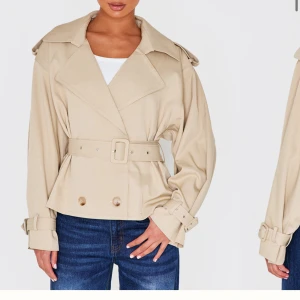 Beige trenchcoat med bälte - Snygg beige trenchcoat med bälte och knappar. Jackan har en klassisk krage och långa ärmar med justerbara spännen vid ärmsluten. Perfekt för en stilren look. Den är även helt ny med lapp på.