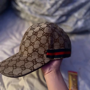 Beige keps helt ny  - Snygg beige keps från shh med det klassiska GG-mönstret i brunt. Kepsen har en justerbar rem baktill och en röd-grön randdetalj på sidan. Perfekt för att ge din outfit en lyxig touch.