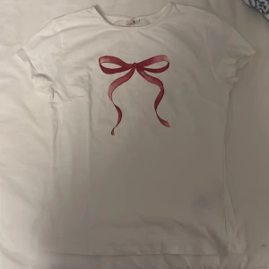 Vit t-shirt med röd rosett - Säljer en söt vit t-shirt med en stor röd rosett tryckt på framsidan från Nelly. 