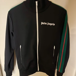 Svart zip-tröja från Palm Angels - Snygg svart zip-tröja från Palm Angels med vit dragkedjar i mycket bra skick. Jackan har långa ärmar med gröna och röda ränder längs sidorna. Nypris: 3000kr (Kvitto finns!)