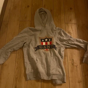 Grå hoodie från Gant - Säljer en snygg grå hoodie från Gant med ett stort tryck på framsidan. Hoodien har en klassisk passform med långa ärmar och en bekväm huva. Perfekt för en avslappnad stil.