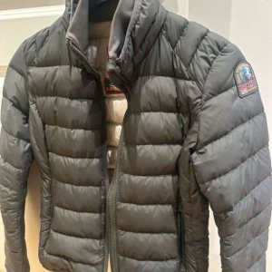 Grå dunjacka från Parajumpers - Säljer en grå dunjacka från Parajumpers med en quiltad design och dragkedja framtill. Jackan har en patch på ärmen och är superlätt, perfekt för kyliga dagar. Insidan är ljus med märkets logga och detaljer.  STORLEK:Young S