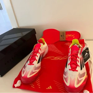Adidas F50 fotbollsskor - Säljer ett par Adidas F50 fotbollsskor i vitt med röda och svarta detaljer. Skorna har snörning och en modern design med en lättviktig konstruktion för optimal hastighet på planen. Perfekta för fotbollsspelare som vill ha både stil och prestanda.