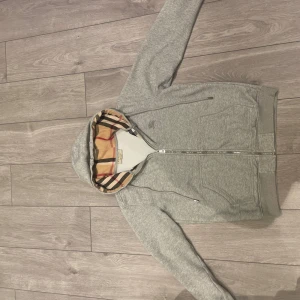 Burberry kofta - Säljer en stilren grå huvtröja från Burberry med dragkedja och klassiskt rutigt mönster i huvan. Tröjan har långa ärmar och en broderad logga på bröstet. Perfekt för en avslappnad och trendig look.