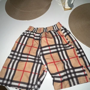 Rutig shorts från Burberry - Snygga b från Burberry med klassiskt rutigt mönster i beige, svart, vit och röd. Shortsen har en elastisk midja och är perfekta för sommardagar vid stranden eller poolen.