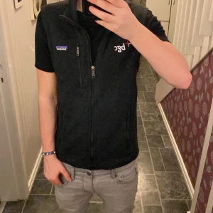 Svart fleeceväst från Patagonia - Säljer en svart fleeceväst från Patagonia med dragkedja och broderad logga. Perfekt för lager-på-lager och håller dig varm under kyliga dagar. Västen har en praktisk bröstficka med dragkedja. Sitter bra på mig som är 170