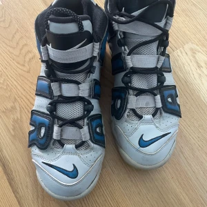 Nike sneakers med blå detaljer - Snygga Nike sneakers i vitt med blå och svarta detaljer. Skorna har en hög design med snörning och det ikoniska Nike-märket på sidan. Perfekta för en sportig look.