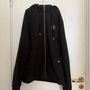 Svart vindjacka från Trapstar - Snygg svart vindjacka från Trapstar med huva och dragkedja framtill. Jackan har ett coolt tryck på ryggen och en rund logga på ärmen. Perfekt för en stilren look oavsett väder.