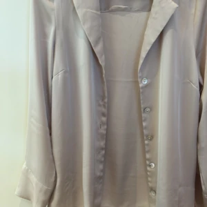 Beige blus från H&M - Elegant beige blus från H&M med knappar framtill. Blusen har en klassisk krage och långa ärmar, perfekt för en stilren look. Materialet ger en mjuk och lyxig känsla.