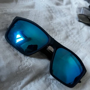 Svarta solglasögon med blå spegelglas från Bliz - Snygga svarta solglasögon med blå spegelglas. Perfekta för soliga dagar och ger en cool look. Ramen är robust och stilren, vilket gör dem till ett måste i sommarens accessoarer.