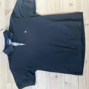 Svart pikétröja från Burberry - Säljer en stilren svart pikétröja från Burberry med klassisk krage och knappslå med rutigt mönster. Tröjan har korta ärmar och diskret broderad logga på bröstet. Perfekt för en avslappnad men elegant look.