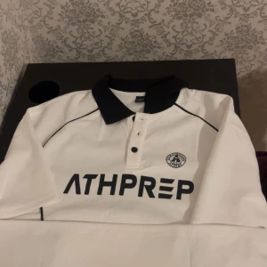 Vit pikétröja från Athprep - Snygg vit pikétröja från Athprep med svart krage och knappar. Tröjan har en stilren design med svart text och logotyp på bröstet. Perfekt för en sportig look.