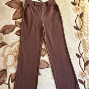 Bruna byxor från First Class Collection - Bruna kostymbyxor från First Class Collection med dragkedja fram. Dragkedjan är dock sönder - därav säljs de för ett billigt pris. De har en klassisk rak passform och är perfekta för en stilren look. Byxorna har hällor för bälte och är tillverkade i ett mjukt och luftigt material. Nypris: 199 kr. Skick: Mycket fint (utom dragkedjan då)