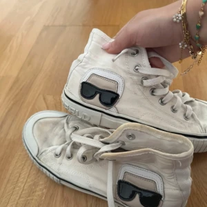 Vita sneakers från Karl Lagerfeld - Snygga vita sneakers från Karl Lagerfeld med coolt svart glasögonmotiv på sidan. Skorna har snörning och en klassisk hög modell. Perfekta för en stilren look med en twist. Skorna finns inte längre att få tag i. Nypris : 1799kr☀️💛🤌🏽