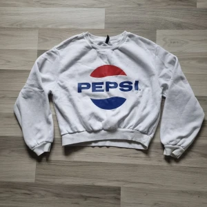 Vit sweatshirt med Pepsi-logga - Säljer en vit sweatshirt från H&M med den klassiska Pepsi-loggan i rött och blått på framsidan. Tröjan har långa ärmar och ribbade muddar vid ärmslut och nederkant. Har en liten röd fläck i mudden i nederkant fram. Perfekt för en avslappnad stil! 🥤