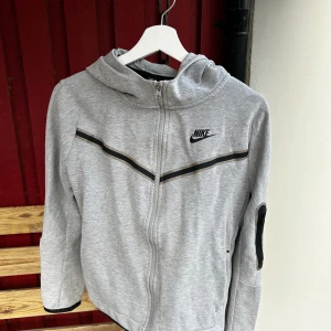 Grå nike tech hoodie - Säljer en grå hoodie från Nike med dragkedja och huva. Tröjan har ett svart Nike-märke på bröstet och svarta detaljer längs ärmarna. Perfekt för en avslappnad stil. Storlek XL i barn storlekar, samma som XS i vuxen storlek.