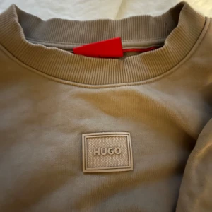 Beige tröja från Hugo - Säljer en stilren beige tröja från Hugo med rund hals och ribbade muddar. Tröjan har en diskret logotyp på bröstet och är perfekt för en avslappnad look. Passar bra till jeans eller chinos.