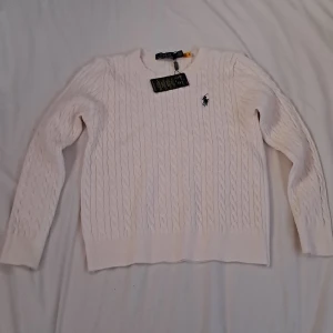 Vit stickad tröja från Polo Ralph Lauren - Säljer en helt ny stilren vit stickad tröja från Polo Ralph Lauren med klassiskt kabelstickat mönster. Tröjan har långa ärmar och en liten broderad logga på bröstet. Perfekt för en elegant och avslappnad look.