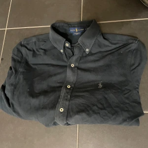 Svart skjorta från Ralph Lauren - Säljer en stilren svart skjorta från Ralph Lauren med broderad logga på bröstet. Skjortan har knappar framtill och en klassisk krage. Perfekt för en elegant look.