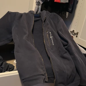 Svart jacka från Sail Racing - Snygg svart jacka från Sail Racing med dragkedja framtill. Jackan har en normal passform och är perfekt för kyligare dagar. Den har långa ärmar och en stilren design med märkets logga på bröstet.