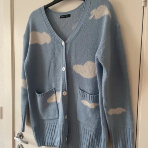 Ljusblå kofta med molnmönster - Säljer en mysig ljusblå kofta med vita moln. Koftan har en v-ringad design med knappar framtill och praktiska fickor. Perfekt för en avslappnad stil.