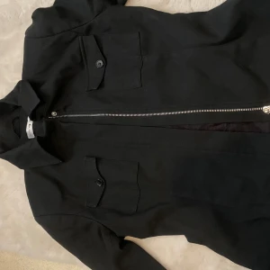 Svart jacka med dragkedja - Snygg svart jacka med dragkedja och knappar på fickorna. Jackan har en klassisk krage och långa ärmar. Perfekt för en stilren look.