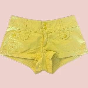 Gula lågmidjade shorts - Snygga gula shorts med låg midja och dubbla knappar fram. De har dekorativa fickor med knappdetaljer och är perfekta för sommardagar. Passar bra till en avslappnad stil.