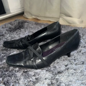 Svarta pumps från Tamaris - Snygga svarta pumps från Tamaris med en elegant klack och spetsig tå. Skorna har dekorativa band över foten som ger en stilren look. Perfekta för en chic och sofistikerad stil.