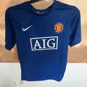 Blå Manchester United fotbollströja - Snygg blå Manchester United fotbollströja från Nike med AIG-logga på framsidan. Tröjan har korta ärmar och klubbens emblem på bröstet. Perfekt för fans som vill visa sitt stöd.