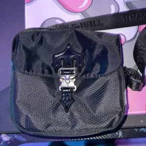 Trapstar - Snygg svart trapstar axelväska med ett unikt spänne på framsidan. Väskan har en justerbar axelrem och är tillverkad i ett slitstarkt syntetmaterial. Perfekt för att bära dina dagliga nödvändigheter med stil.