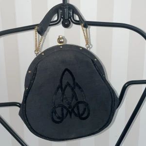 Svart handväska med guldkedja - Elegant svart handväska med en unik form och dekorativt mönster på framsidan. Mått 20*20 cm utan handtag.Väskan har en guldkedja och en praktisk bygelöppning med guldfärgad ram. Perfekt för att ge en stilfull touch till din outfit.