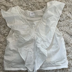Vit ärmlös topp från Zara - Säljer en söt vit ärmlös topp från Zara med volangdetaljer framtill. Perfekt för varma sommardagar. Toppen är tillverkad i ett lätt och luftigt material.