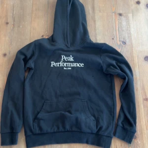 Svart hoodie från Peak Performance - Säljer en svart hoodie från Peak Performance med vit logga på bröstet. Tröjan har en klassisk känguruficka och ribbade muddar vid ärmslut och nederkant. Perfekt för en avslappnad stil.