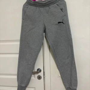 Grå mjukisbyxor från Puma - Säljer ett par grå mjukisbyxor från Puma med en klassisk logga på sidan. Byxorna har en bekväm passform och är perfekta för en avslappnad stil. De är tillverkade i mjukt material för extra komfort.