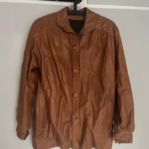 Brun retro skinnjacka med knappar - Vintage brun skinnjacka med knappar framtill. Jackan har långa ärmar och en klassisk krage. Retro jacka tillverkad i Malung av Pg Elfström. Storlek Herr 36, passar dam M. Skav finns på lädret.