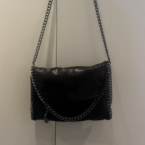 Svart axelväska med kedjedetaljer - Snygg svart axelväska med glittrig yta och kedjedetaljer. Väskan har ett leopardmönstrat foder och en praktisk dragkedja inuti.